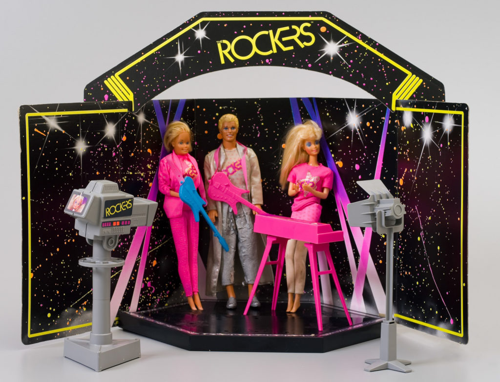 The two Barbie dolls' costumes consist of a pink 'Rockers' T-shirt, one doll has a pink mini-skirt and tan leggings; the other has a pink jacket and pants ensemble. The styling is indicative of the mid 1980s. The Ken doll's costume is a silver long jacket and pants ensemble with a silver 'Rockers' T-shirt, and silver shoes.